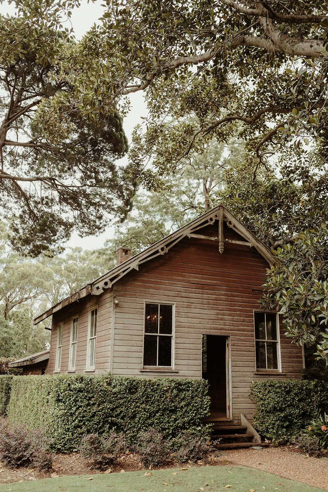 A rustic wooden house with a gabled roof, surrounded by lush greenery and trees. The house features multiple windows and an open door. Tall hedges and a lawn are in the foreground, and the overall scene looks peaceful and serene.