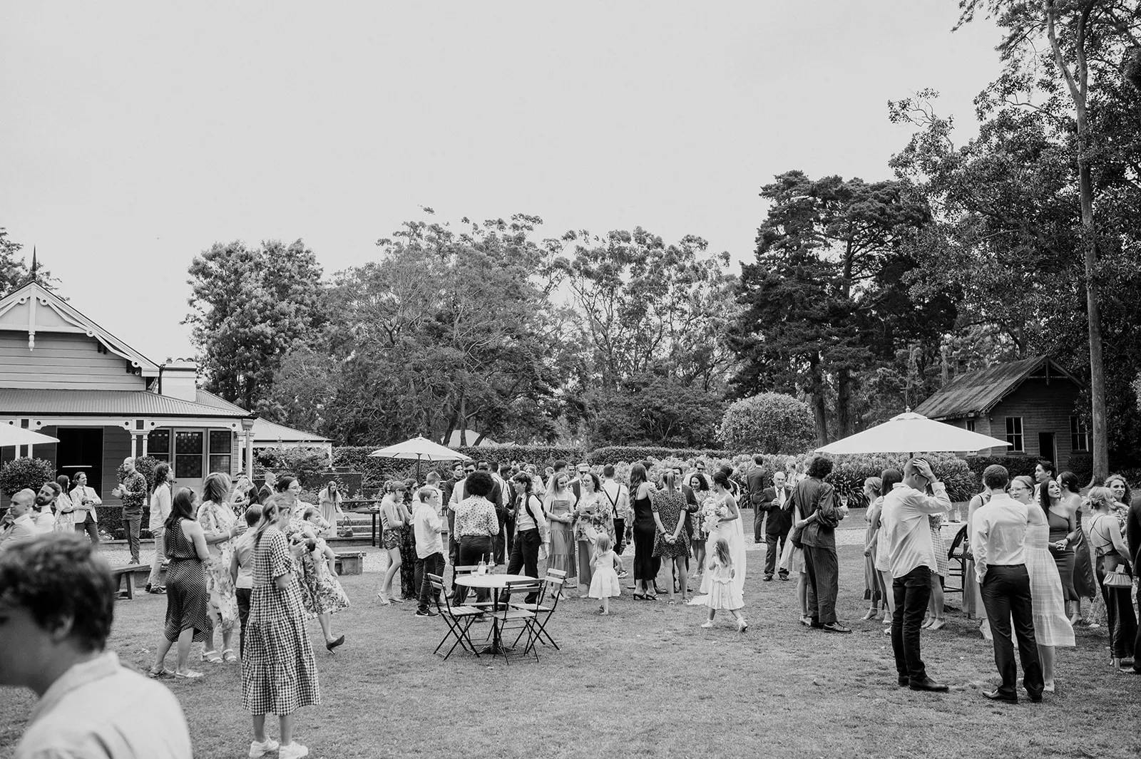 Black and white photo of an outdoor gathering in a garden. People of various ages are standing and interacting near small tables and chairs. Trees and a house are visible in the background. Some attendees are holding glasses, possibly at a celebratory event.