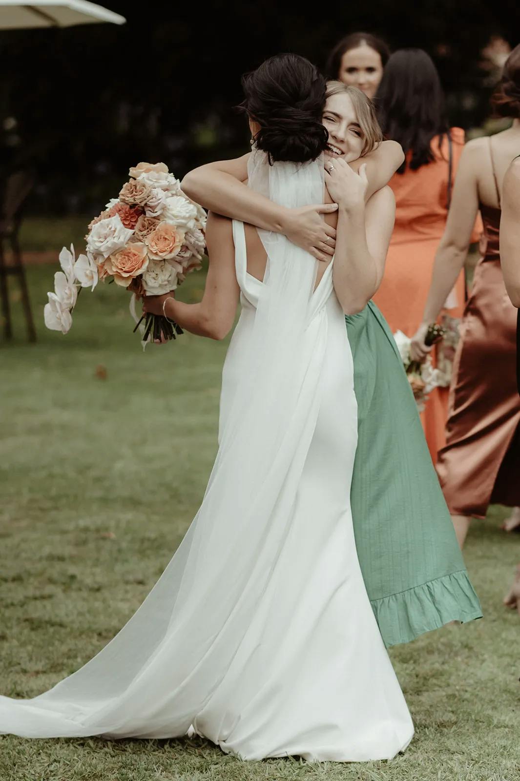 A bride in a white dress holding a bouquet of flowers hugs a woman in a green dress at an outdoor event. Other people dressed in formal attire are seen in the background. The atmosphere appears joyous and celebratory.