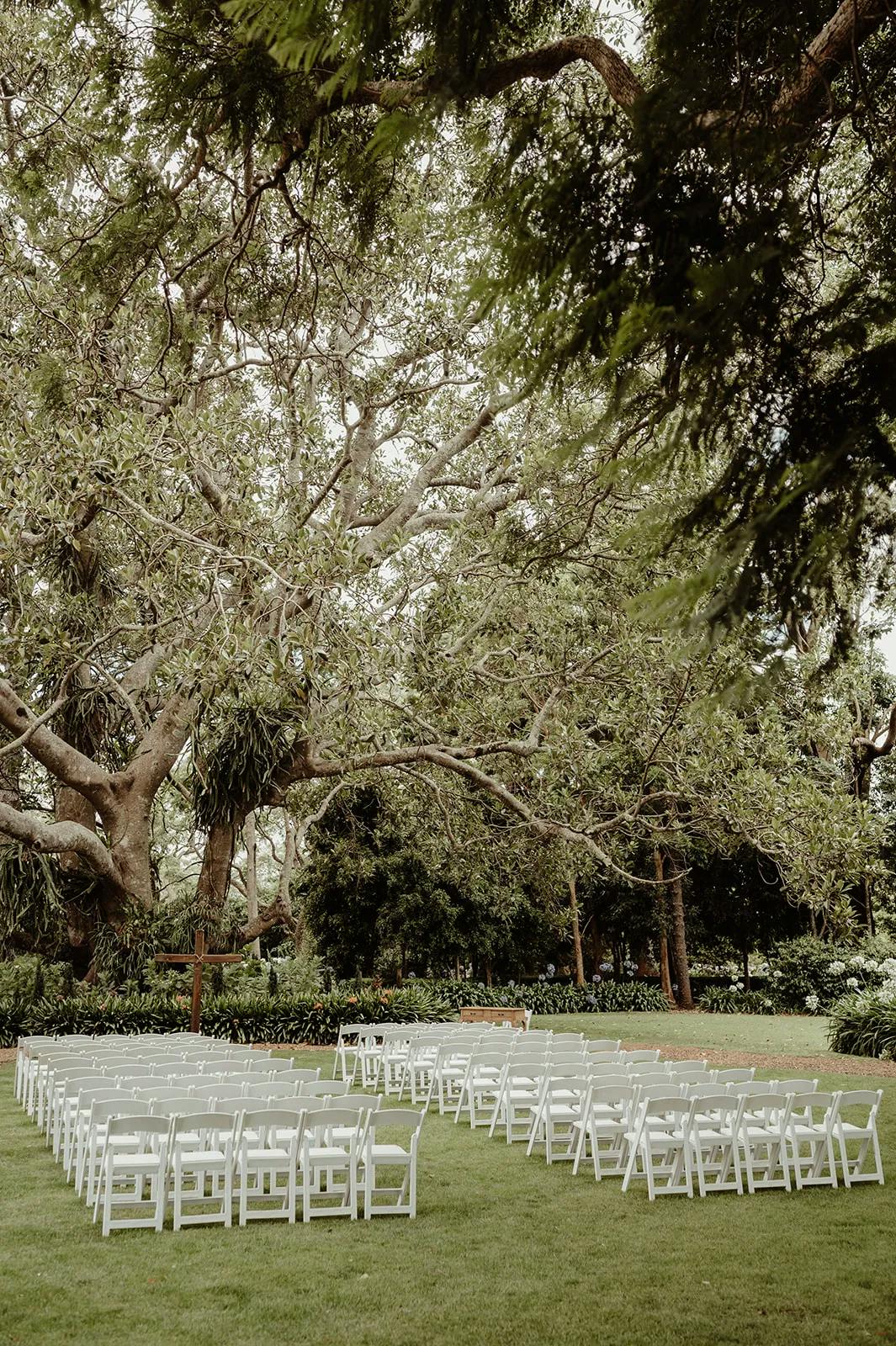 An outdoor wedding setup featuring rows of white chairs arranged in a neatly organized rectangular formation on a lush green lawn. The area is surrounded by large, leafy trees creating a serene and picturesque backdrop.