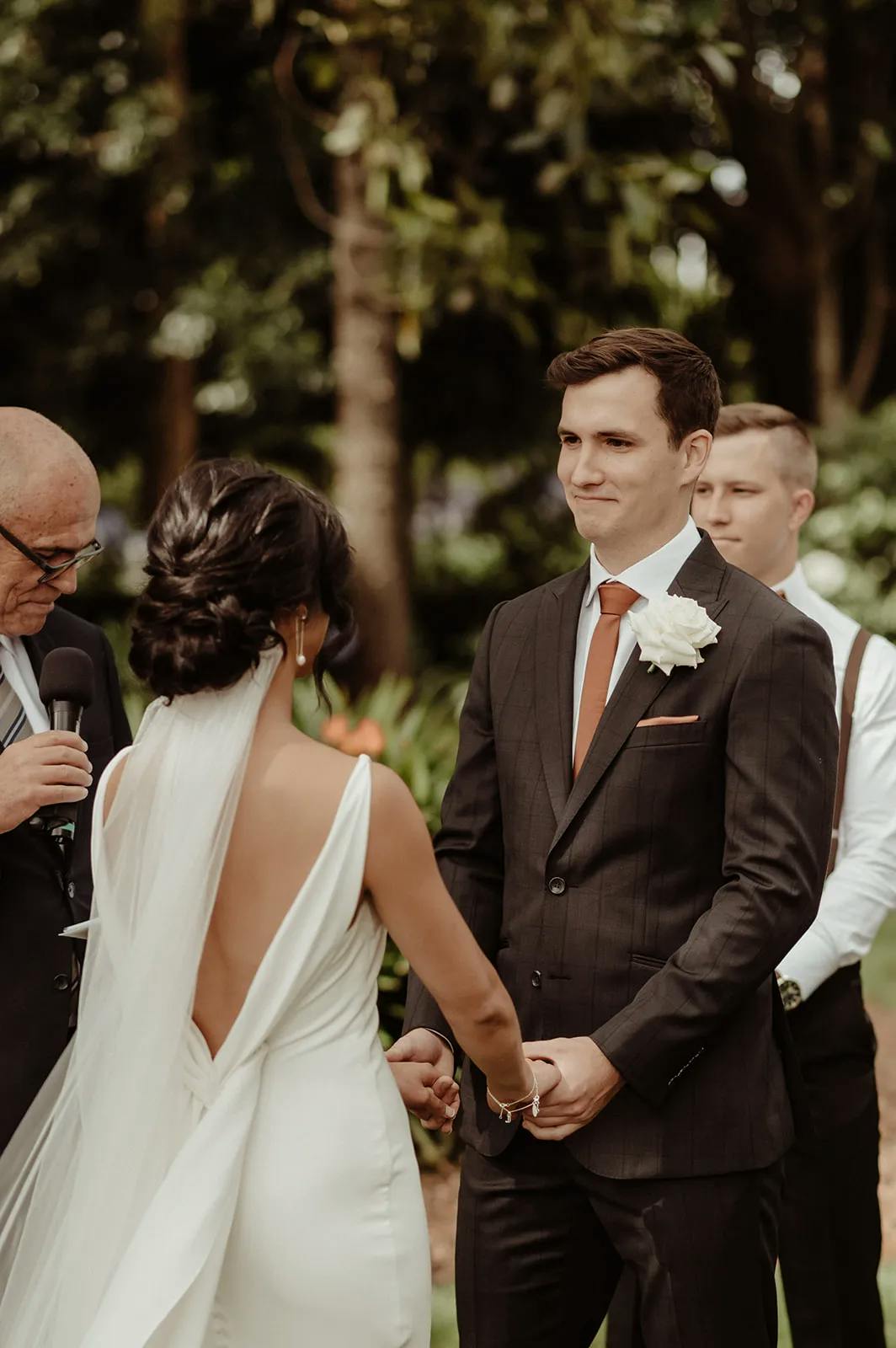 A bride and groom stand facing each other, holding hands, during their outdoor wedding ceremony. The groom wears a dark suit with a white flower boutonniere, while the bride wears a white dress with a deep back and a long veil. A person officiates the ceremony.