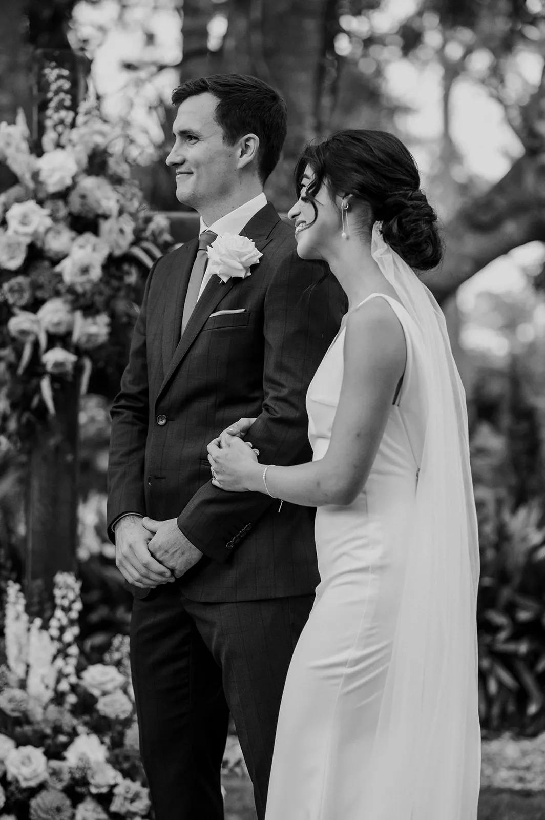 A bride and groom stand together, smiling and looking in the same direction. The bride, in a white gown with a long veil, holds the groom's arm. The groom wears a dark suit with a white rose boutonniere. They are outdoors near a floral arrangement. The photo is in black and white.