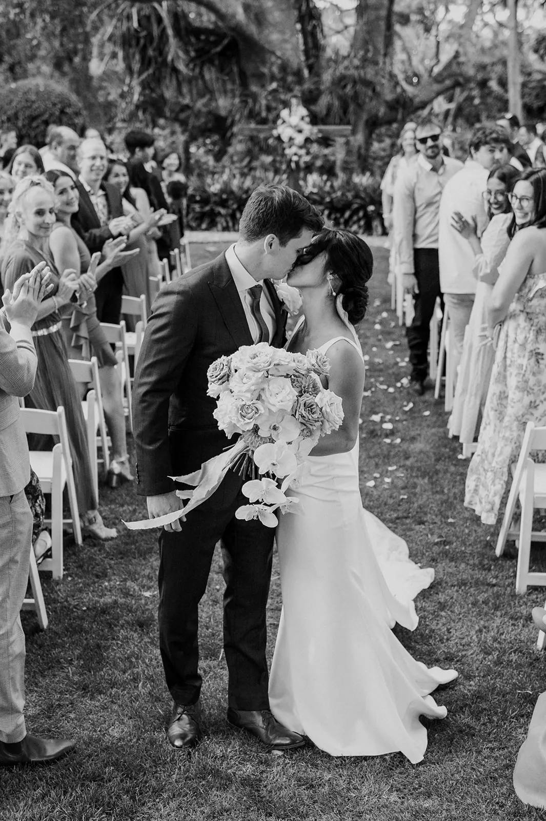 A couple shares a kiss while holding a bouquet of flowers as they walk down the aisle at their outdoor wedding ceremony. Guests on both sides stand and applaud. The setting is lush and green, suggesting a garden venue. The photo is in black and white.