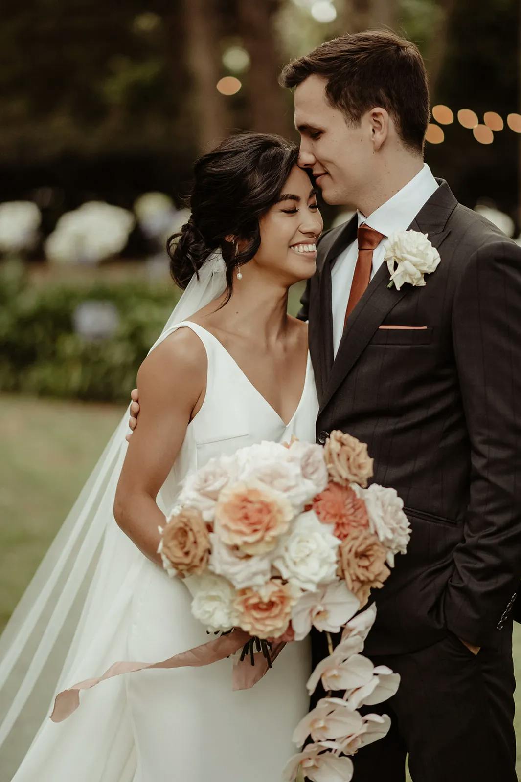 A bride and groom share a loving moment outdoors. The bride, in a white dress with a veil, holds a bouquet of flowers and smiles with closed eyes, while the groom, in a dark suit with an orange tie, gently leans his forehead against hers. The background is lush and green.