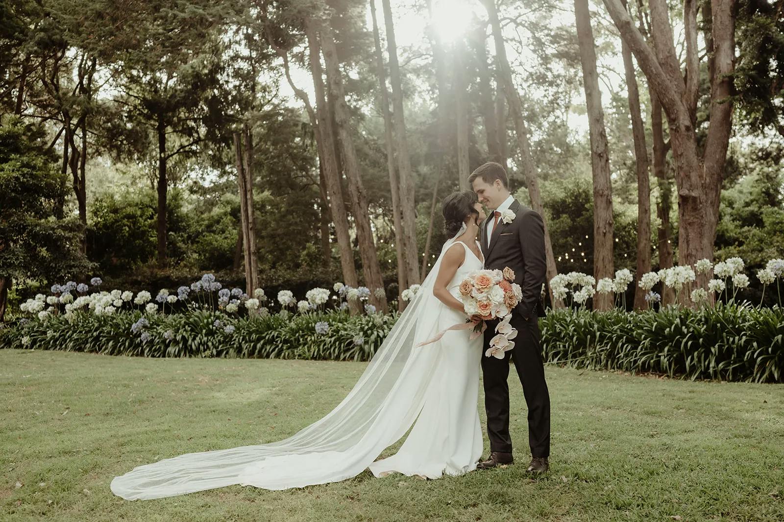 A bride and groom stand facing each other in a lush garden with tall trees and flower bushes. The bride, in a white gown with a long train, holds a bouquet of pink flowers. The groom, in a black suit, gazes at her lovingly. Sunlight filters through the trees behind them.