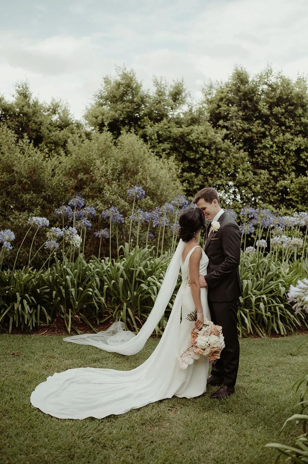 A bride and groom stand together in a lush garden, surrounded by greenery and purple flowers. The bride wears a long white dress with a flowing train and veil, holding a bouquet of flowers. The groom is dressed in a black suit. They gaze into each other's eyes lovingly.