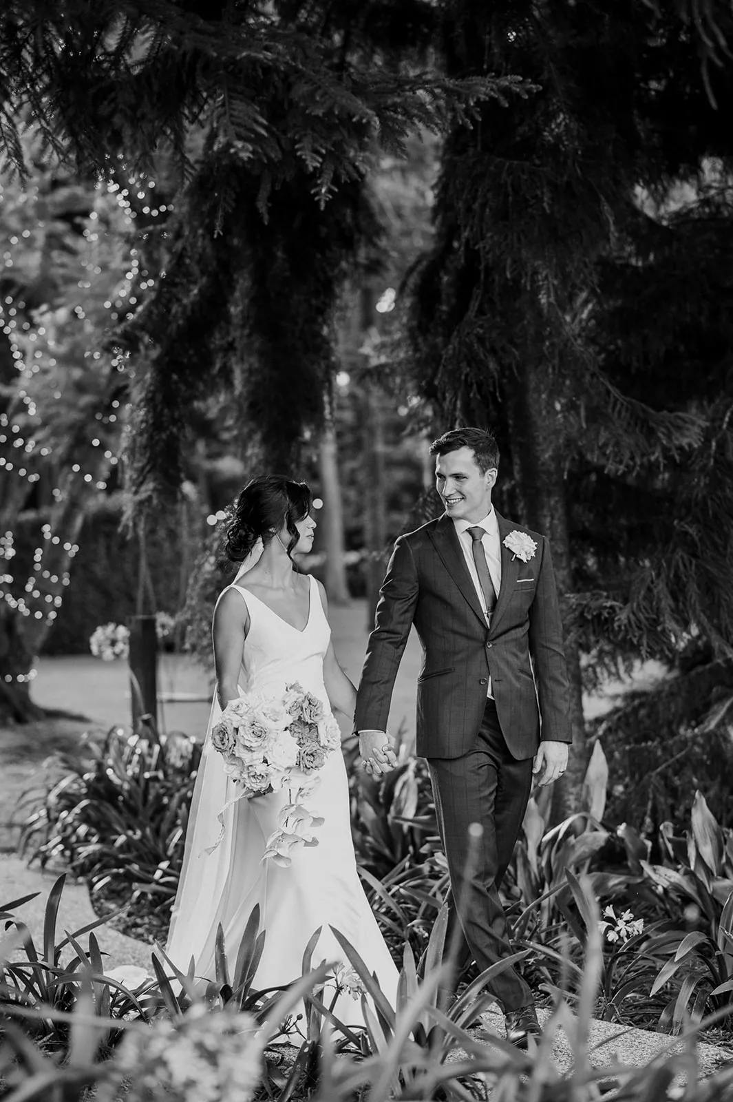 Black and white photo of a bride and groom walking hand in hand in an outdoor setting. The bride is wearing a white dress and holding a bouquet, while the groom is wearing a dark suit with a boutonnière. They are surrounded by trees and foliage, with lights in the background.