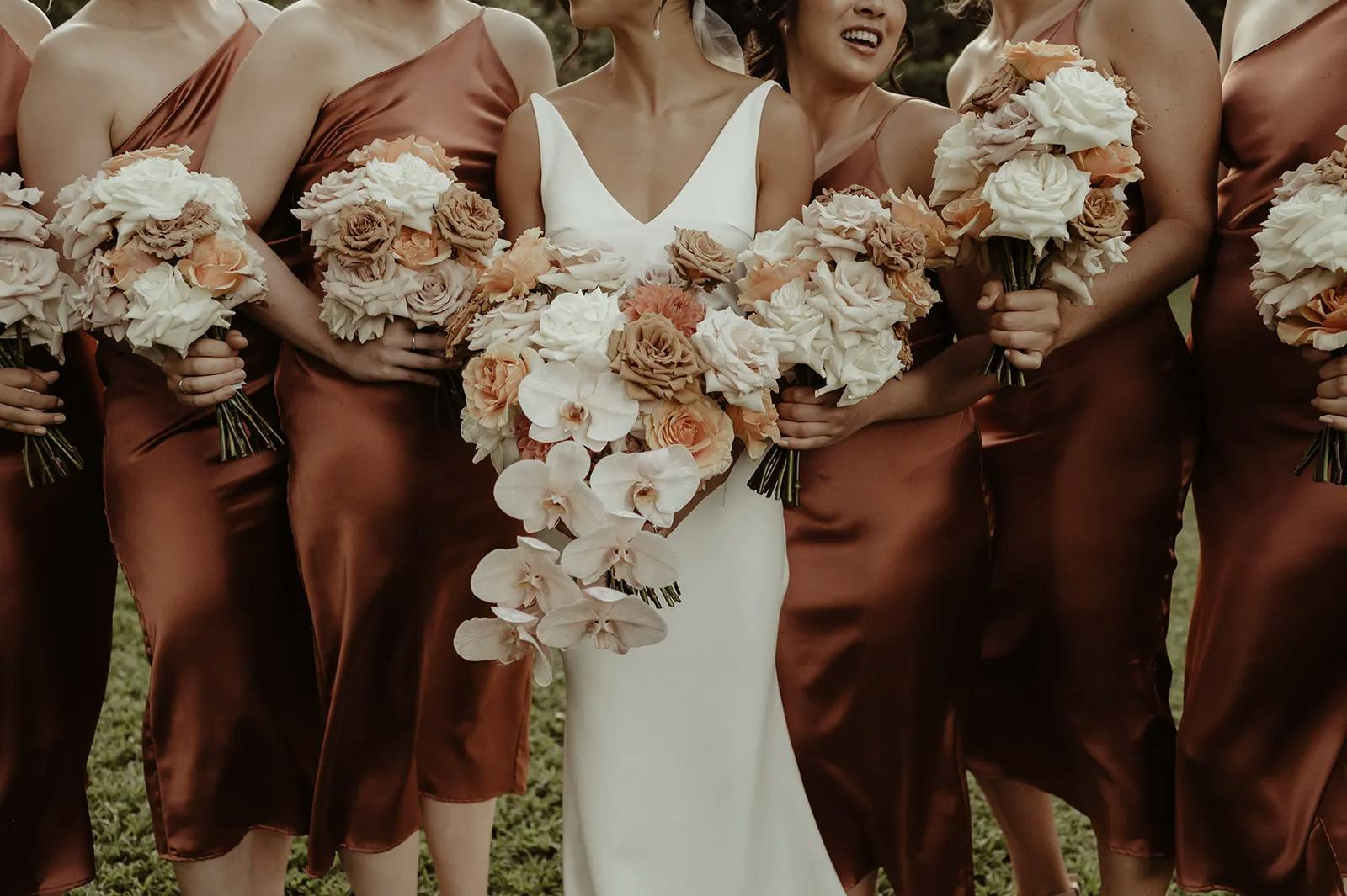 A group of bridesmaids in matching silky bronze dresses stands in a line, each holding a bouquet of white and peach flowers. The bride in the center wears a white gown and holds a large bouquet of white and peach flowers, including orchids.