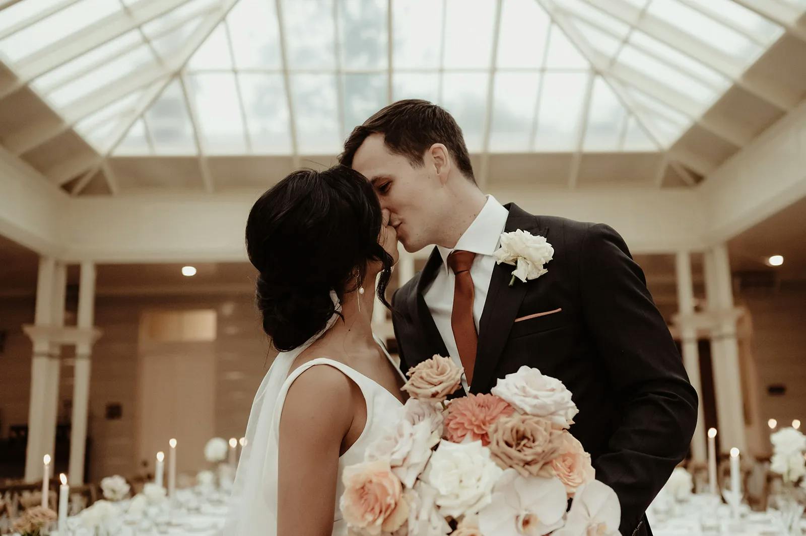A bride and groom share a kiss under a glass-ceilinged venue adorned with white floral arrangements and candles. The bride holds a bouquet of pastel-colored roses, and both are dressed in formal attire with the groom in a dark suit and the bride in a sleeveless gown.