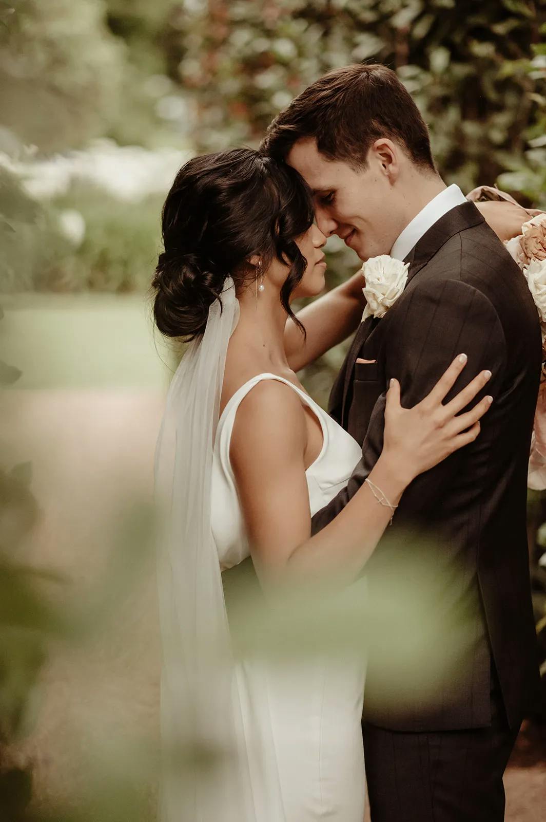 A couple in wedding attire embraces tenderly in a garden setting. The bride, in a white dress and veil, and the groom, in a dark suit and tie, touch foreheads and smile softly. They are surrounded by greenery, creating an intimate and romantic atmosphere.
