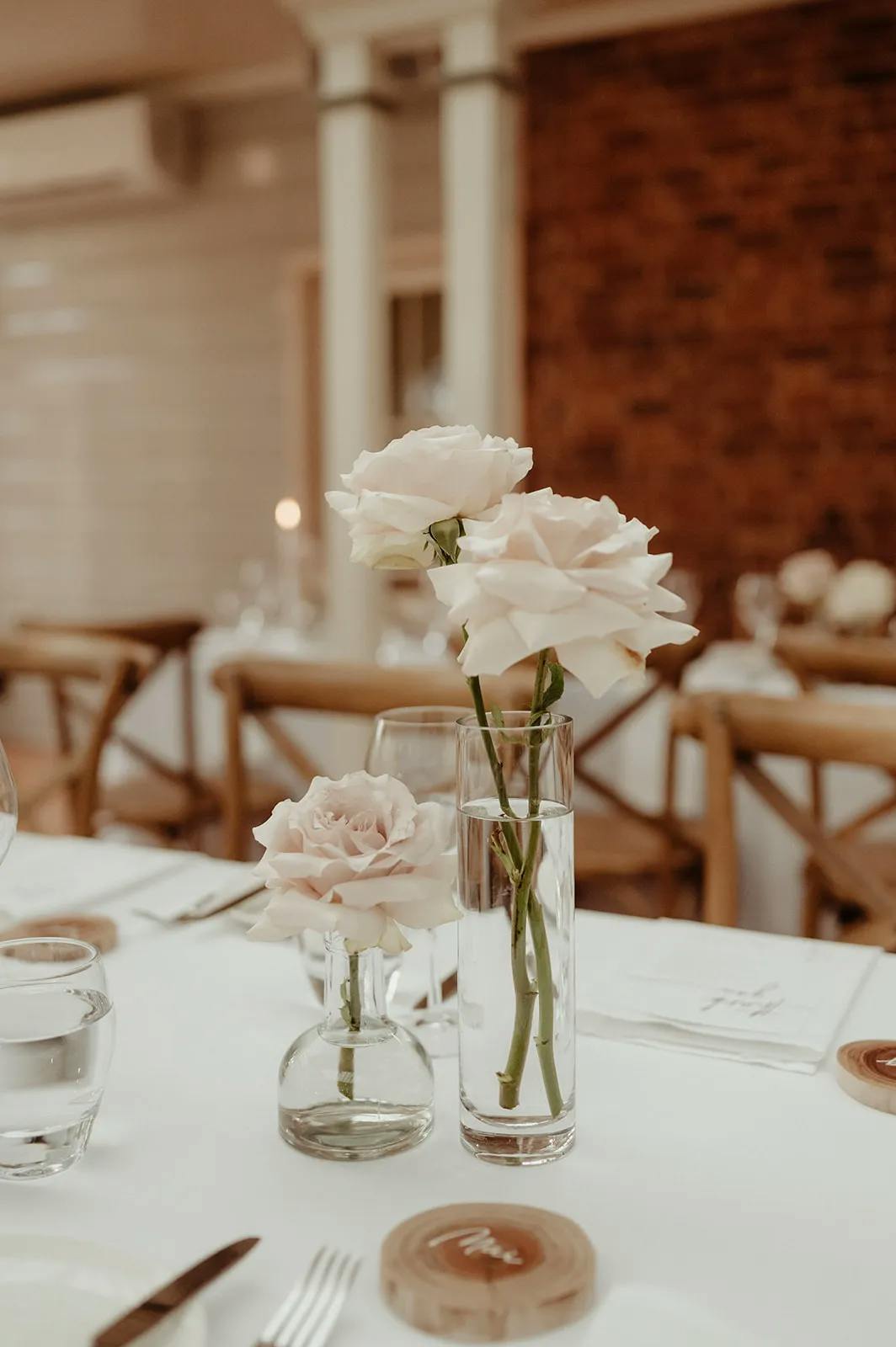 A table decorated with two vases holding light pink roses, one taller and one shorter. The table is set with glassware, white plates, and small wooden place markers. The background features wooden chairs and a mix of brick and light-colored walls.