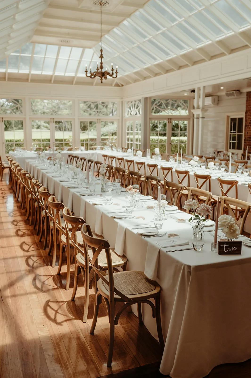 A sunlit elegant banquet hall with long, white-clothed tables set for a formal event. Wooden chairs line the tables, each adorned with plates, glasses, and simple floral centerpieces. The room features large windows and a high, glass-paneled ceiling.