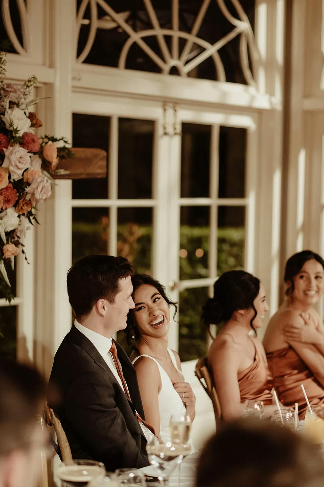 A bride and groom sit smiling at their wedding reception table. The bride, in a white dress, looks joyfully at the groom, who wears a suit and tie. Other guests, also smiling, are seated beside them. The background features large windows and a floral arrangement.