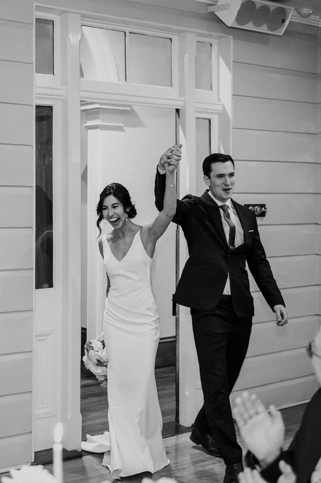 A newlywed couple holds hands and smiles joyfully as they walk into a room. The bride is in a white gown holding a bouquet, and the groom is in a dark suit and tie. The room has wooden walls and people are seen clapping in the foreground. The image is in black and white.
