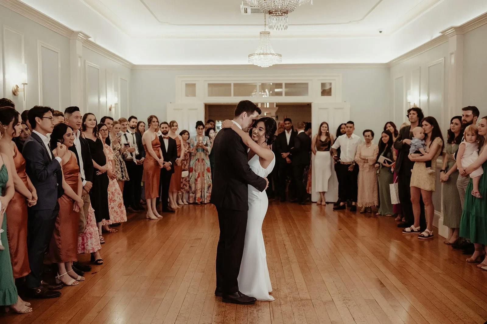 A bride and groom share their first dance in the center of a ballroom, surrounded by guests who watch and smile. The room is brightly lit with chandeliers, and the crowd includes people in formal and semi-formal attire, standing along the walls.