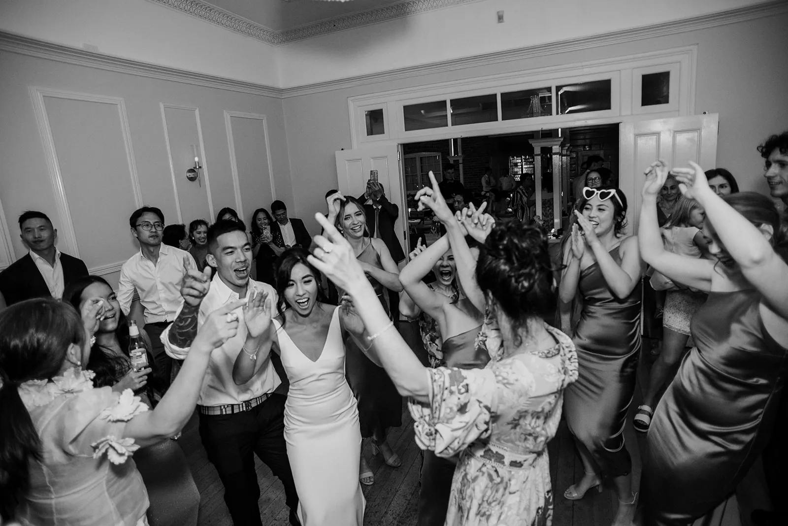 A black and white photo of a wedding reception where a group of people, including the bride and groom, are joyfully dancing together in a circle. The guests are smiling, raising their hands, and embracing the celebratory atmosphere in a decorated room.