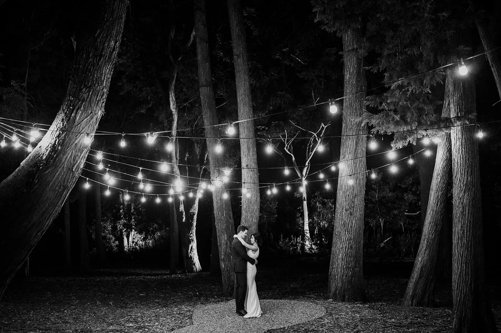 A couple embraces under a canopy of string lights in a forest at night. The warm lights contrast with the dark trees, creating a romantic and intimate atmosphere. The bride is in a long dress, and the groom is in a suit.