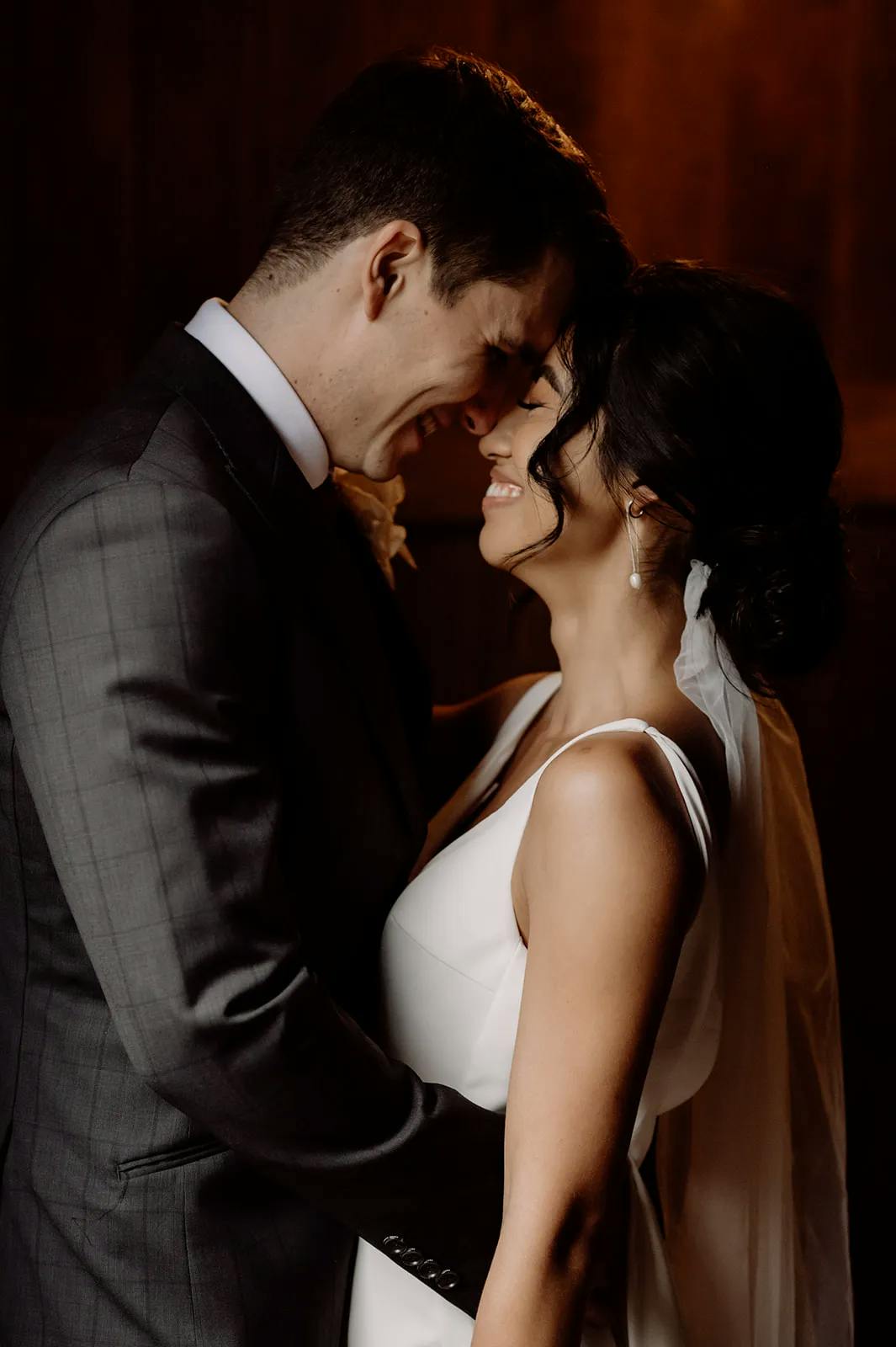 A bride and groom stand closely, forehead to forehead, smiling and basking in a warm light. The bride wears a white dress with a veil and has her hair styled in a low bun. The groom is dressed in a dark suit with a white dress shirt.