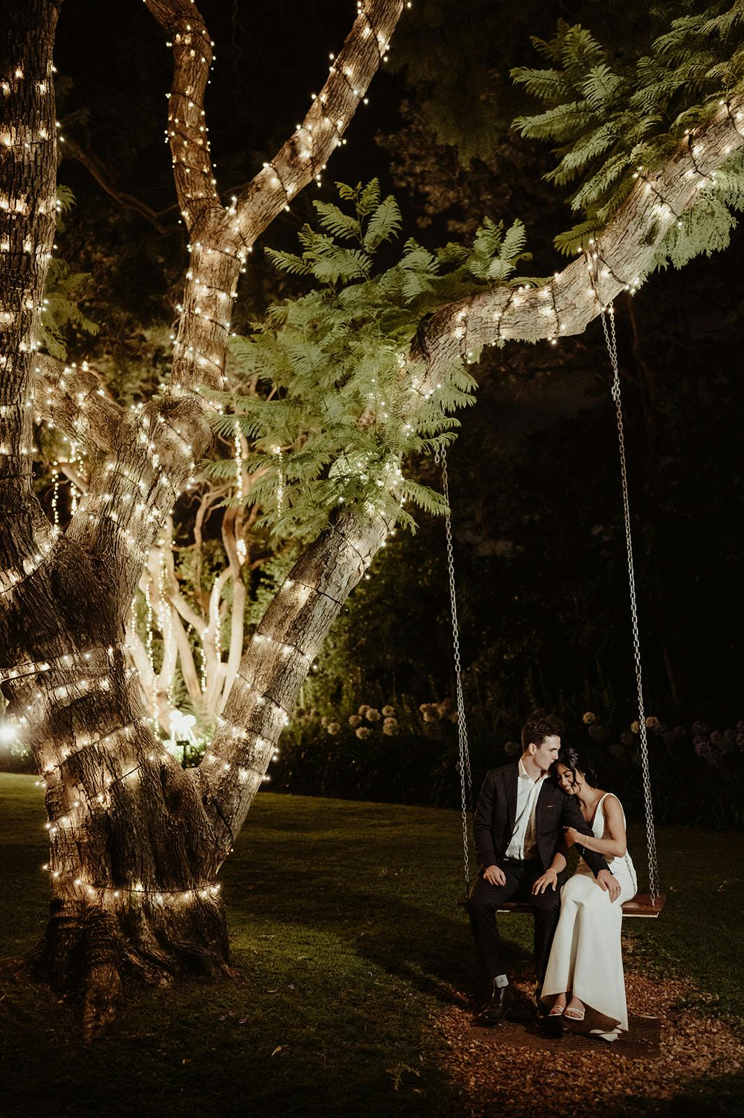 A couple dressed formally sit closely on a swing, suspended from a large tree wrapped in string lights, creating a romantic and serene night-time atmosphere. The woman lays her head on the man's shoulder as they sit under the illuminated branches.
