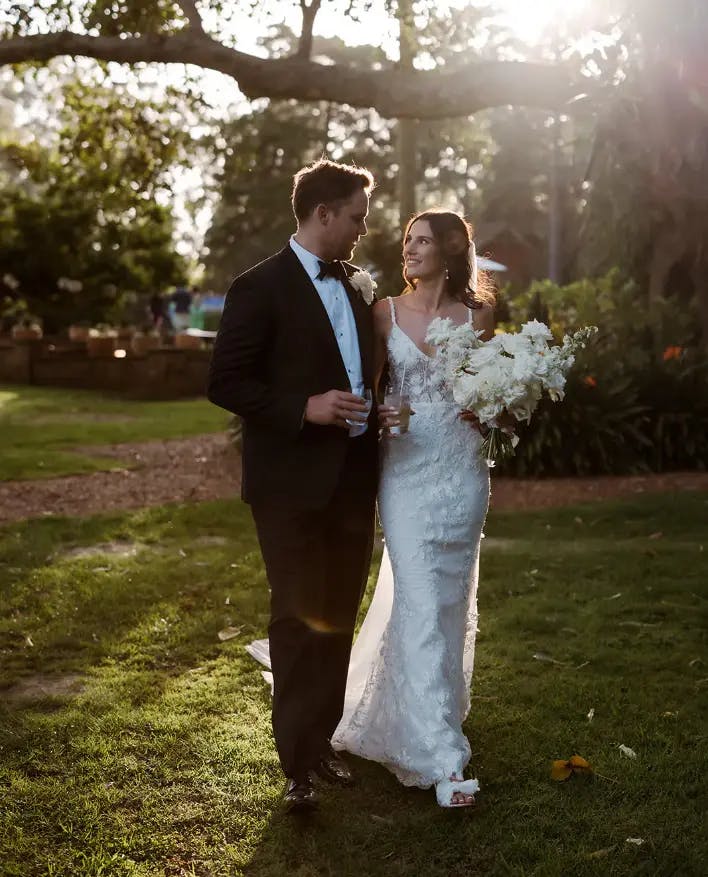 A couple walks outdoors on a grassy path during their wedding. The groom is dressed in a black tuxedo, while the bride wears a white lace dress and holds a bouquet of white flowers. They are looking at each other affectionately with sunlight streaming through the trees.