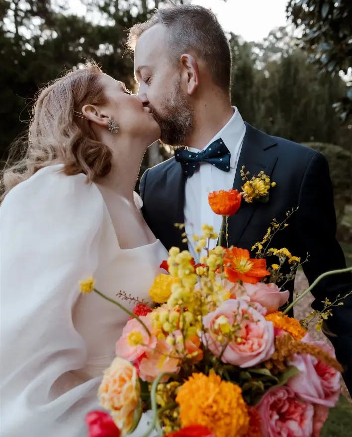 A bride and groom share a kiss on their wedding day. The bride is holding a vibrant bouquet with pink, yellow, and orange flowers, and she is wearing a white dress. The groom is in a dark suit with a bow tie. They are outdoors with greenery in the background.