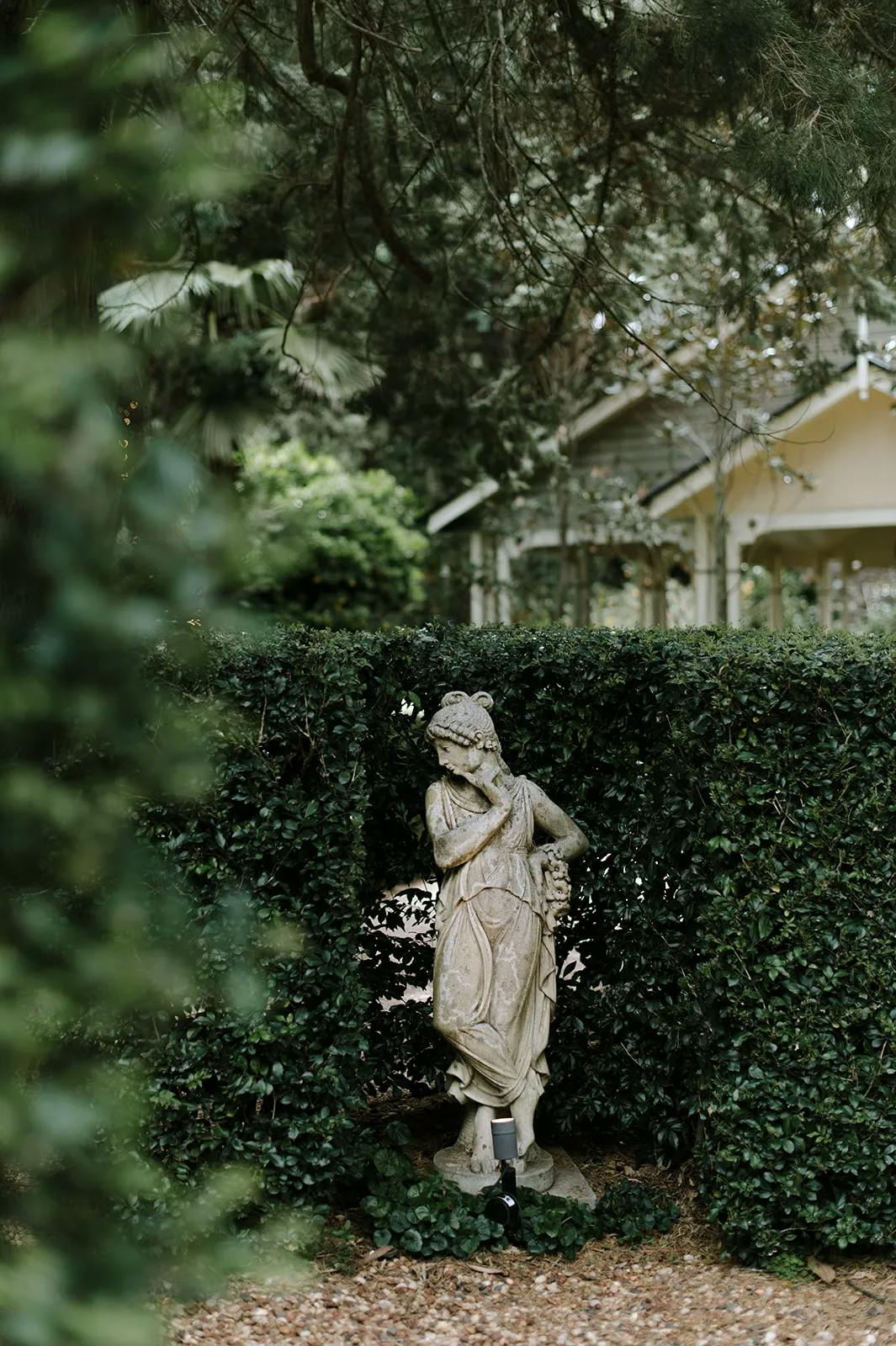 A stone statue of a woman in classical attire stands partially obscured by dense green bushes and surrounded by tall trees. In the background, a light-colored building with a porch is visible through the foliage, adding to the serene garden ambiance.