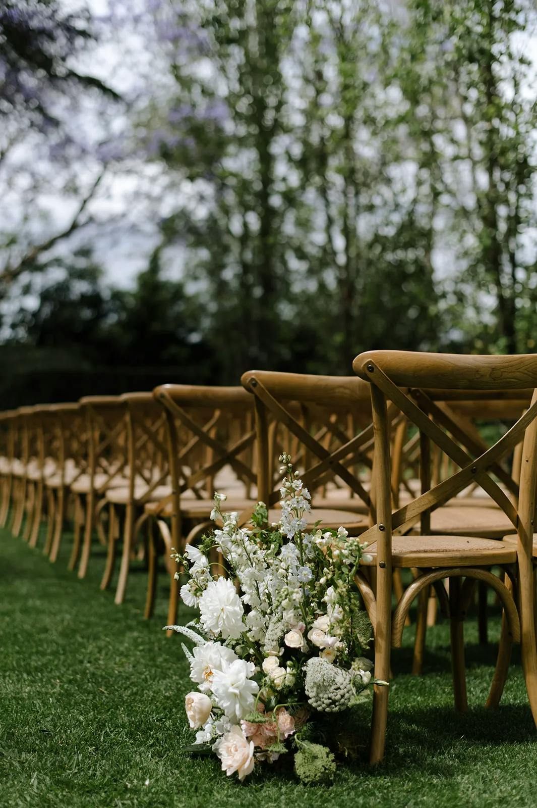 A row of wooden chairs with crossed backs is set up outdoors on a grassy lawn. The chairs are aligned neatly, and the area is decorated with white and green floral arrangements. Trees and a purple-hued sky can be seen in the background, giving a serene atmosphere.
