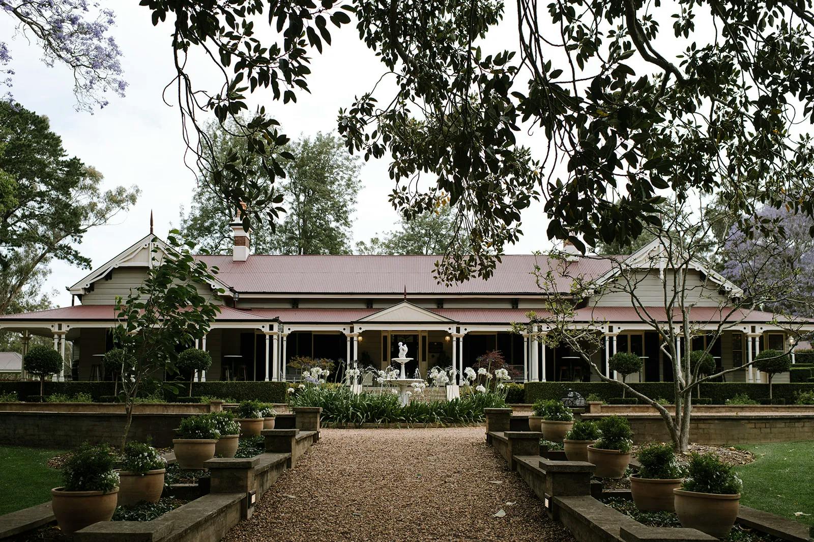 A charming, one-story farmhouse with a red roof and a wide porch, framed by hanging branches and potted plants along a gravel path. The garden features a white fountain surrounded by green foliage and flowers, enhancing the serene countryside setting.