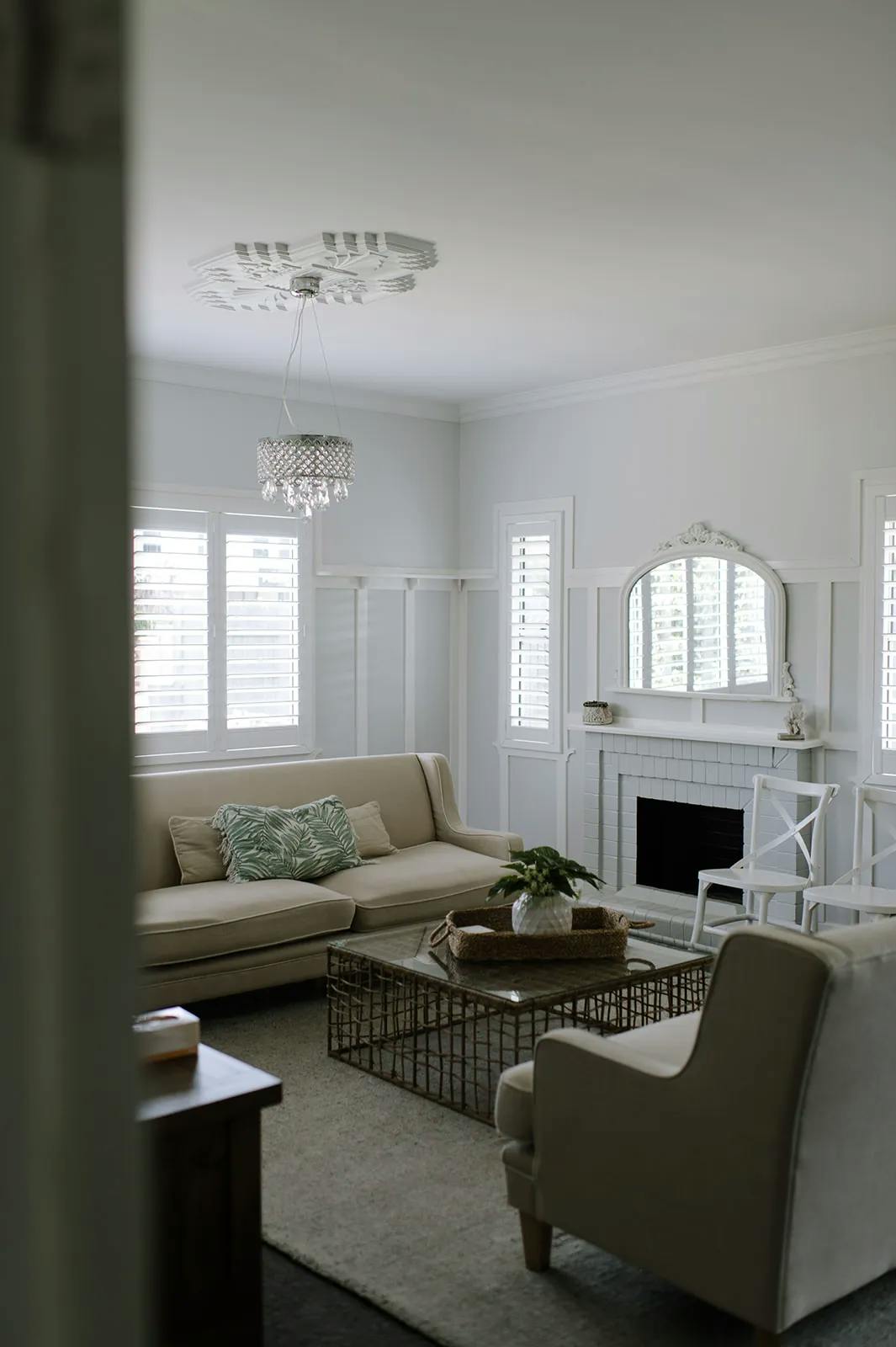 A cozy living room with white walls and a large window with white shutters. The room features a beige sofa, a white armchair, a glass coffee table with a plant, a fireplace with a mirror above it, and a decorative chandelier on the ceiling.