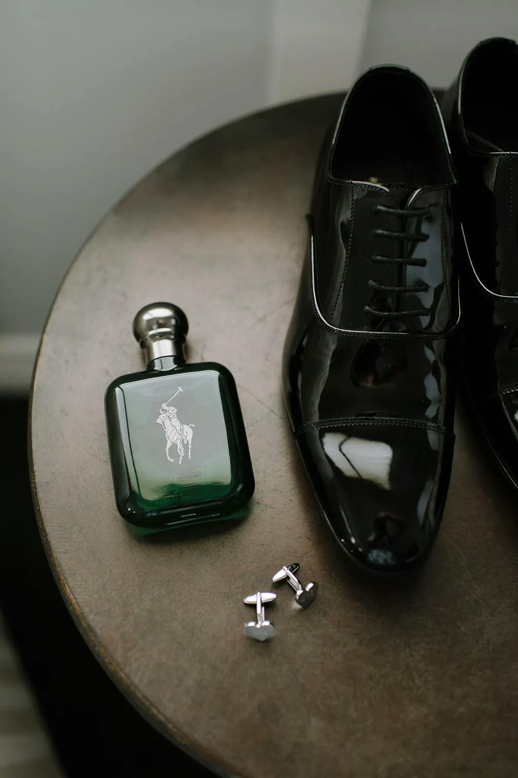 A pair of polished black dress shoes, a green glass bottle of Ralph Lauren cologne with a silver cap, and a set of silver cufflinks are neatly arranged on a round wooden table. The cufflinks feature a minimalist design with straight edges.