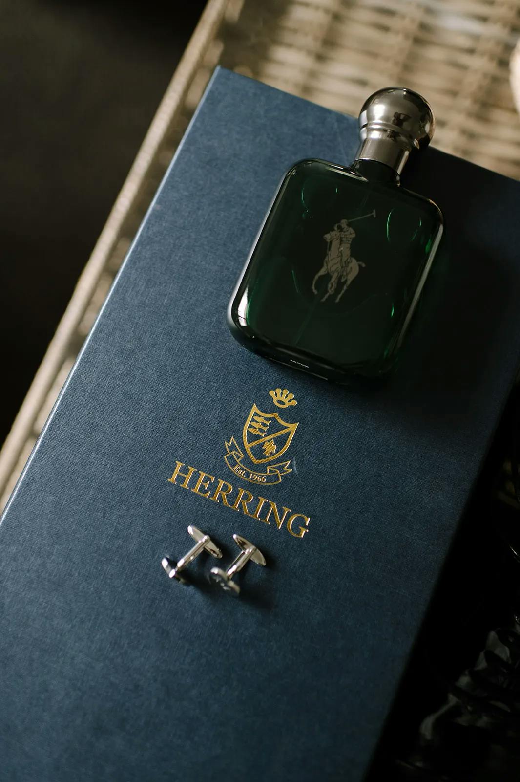 A dark green glass bottle of cologne with a silver cap rests on a dark blue box labeled "Herring" with a gold crest. Beside the cologne, a pair of silver cufflinks shaped like the letter "H" are placed on the box.