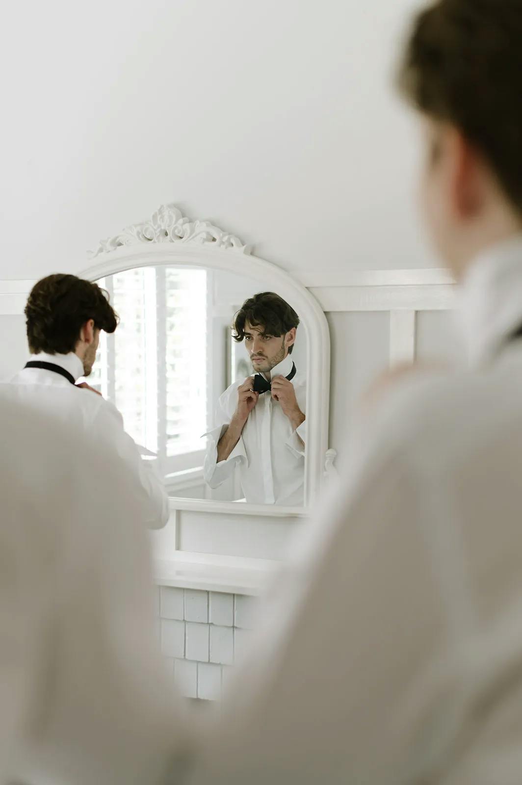 A man in formal attire adjusts his bow tie while looking in a white-framed mirror. The reflection shows his focused expression. The background features white walls and a window with blinds, creating a bright and elegant setting.