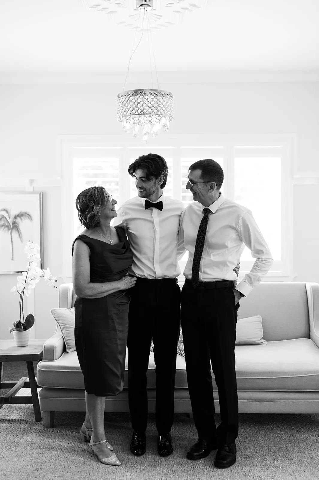 A black and white photo showing three people smiling and standing in front of a couch. The person in the middle is wearing a bow tie and suit, and is flanked by two older individuals, one on each side, each with an arm around the person's shoulders. A chandelier hangs above.