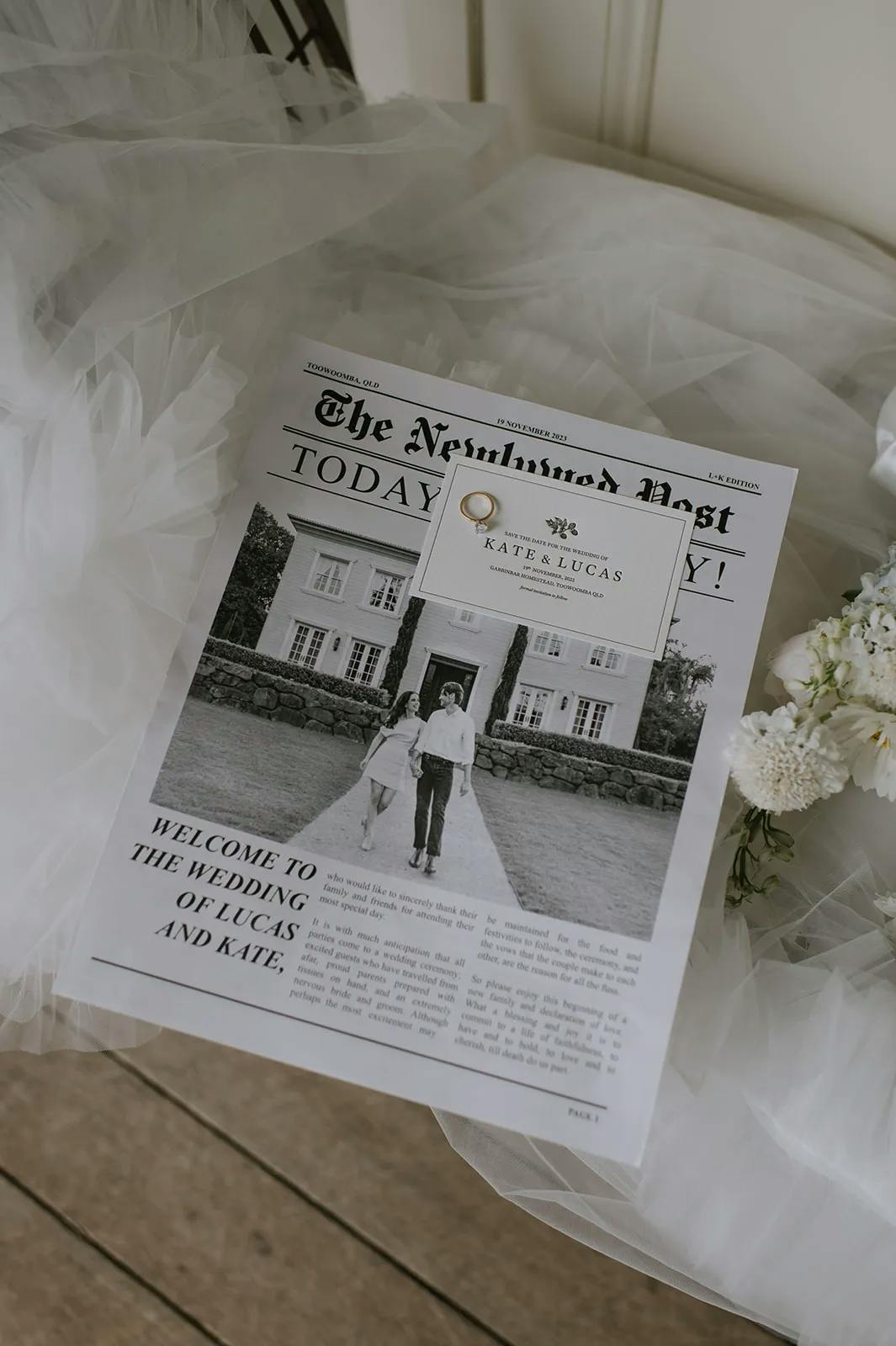 A wedding program styled as a newspaper titled "The Newlywed Post" rests on a veil beside white flowers. The cover features a black-and-white photo of a couple holding hands in front of a house. The headline reads "Welcome to the Wedding of Lucas and Kate.