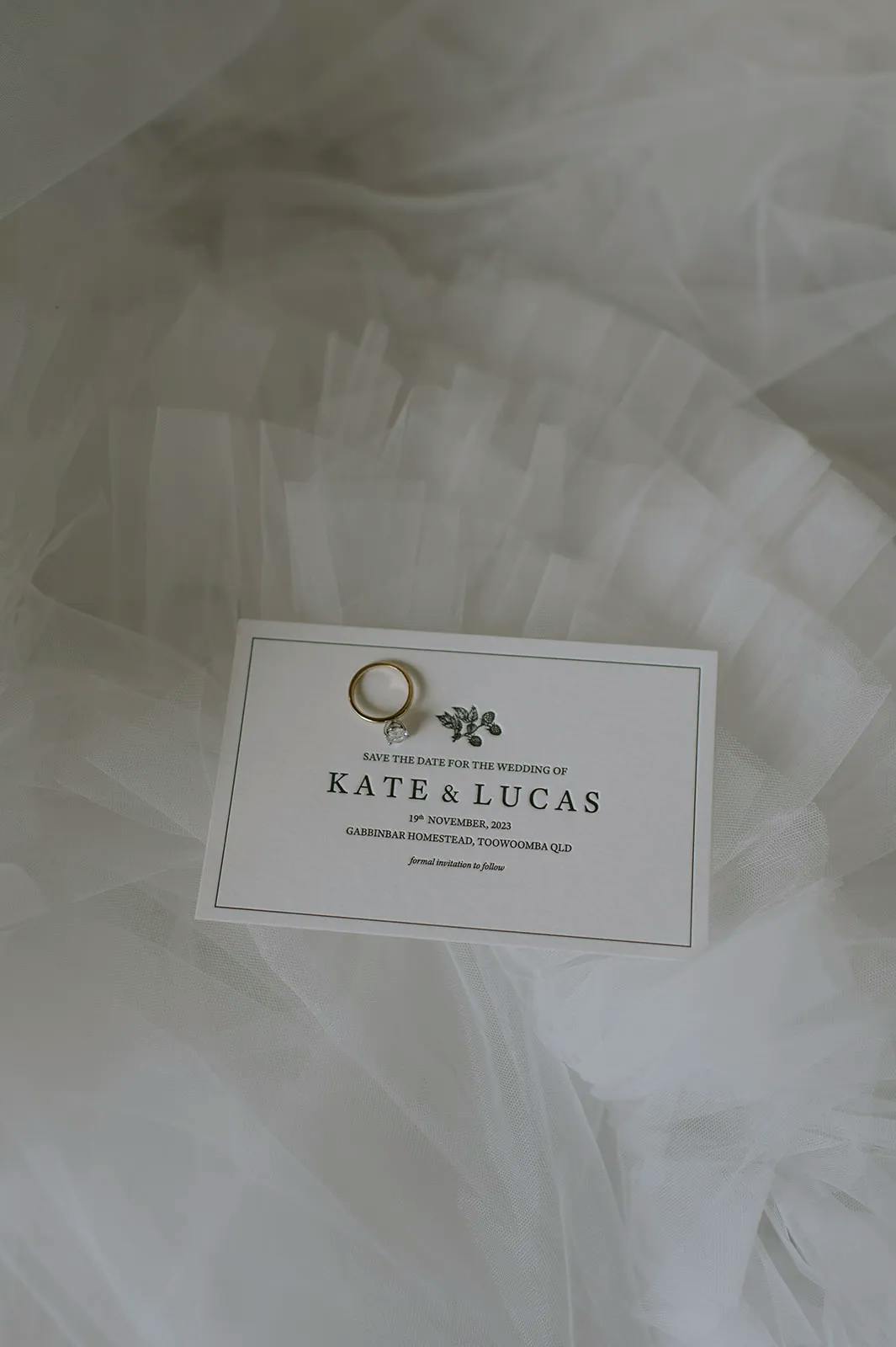 A gold wedding ring is placed on a "Save the Date" card. The card, set on a soft, flowing tulle background, announces the wedding of Kate and Lucas on 18 November 2023 at Gabbinbar Homestead, Toowoomba, QLD.
