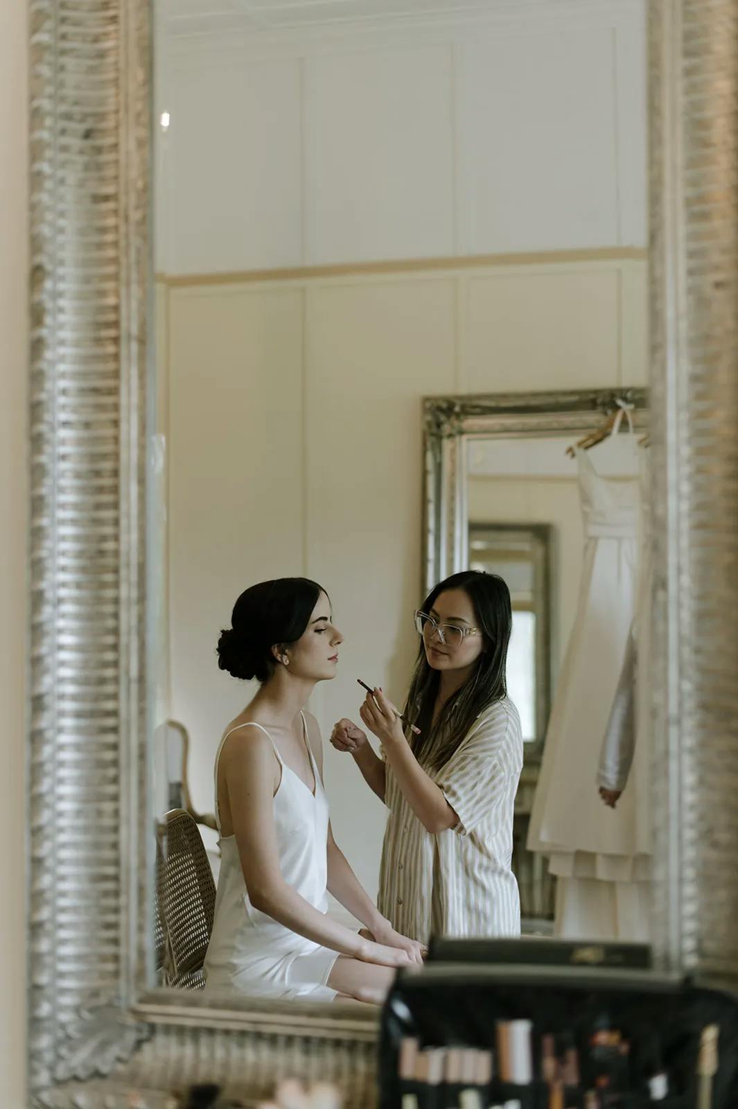 A woman wearing a white dress sits on a chair in front of a large mirror, while another woman, standing, applies makeup to her face. A white wedding dress hangs on the wall in the background. Various makeup tools are seen in the foreground.
