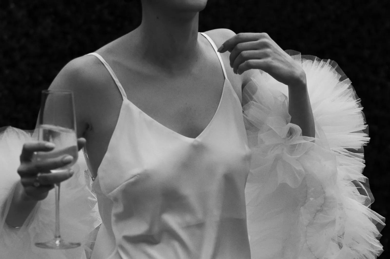 A black and white photograph of a person wearing a delicate, strappy dress with ruffled tulle sleeves. The person is holding a champagne flute in their right hand, and their left hand is raised, touching their shoulder. The background is dark and out of focus.