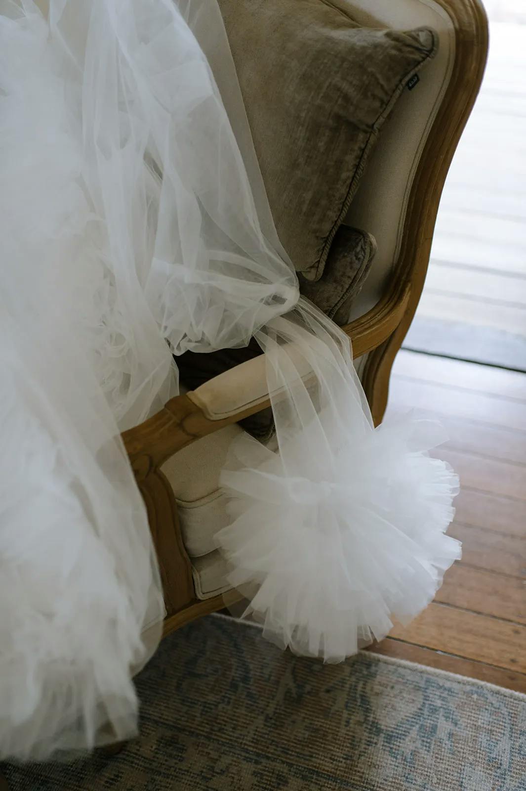 A close-up of a luxurious, vintage-style armchair with beige cushions and wooden frame. A white tulle veil or gown is draped over the chair, with a small pouf of tulle hanging off the arm, adding a soft, elegant touch to the scene. The floor is wooden.