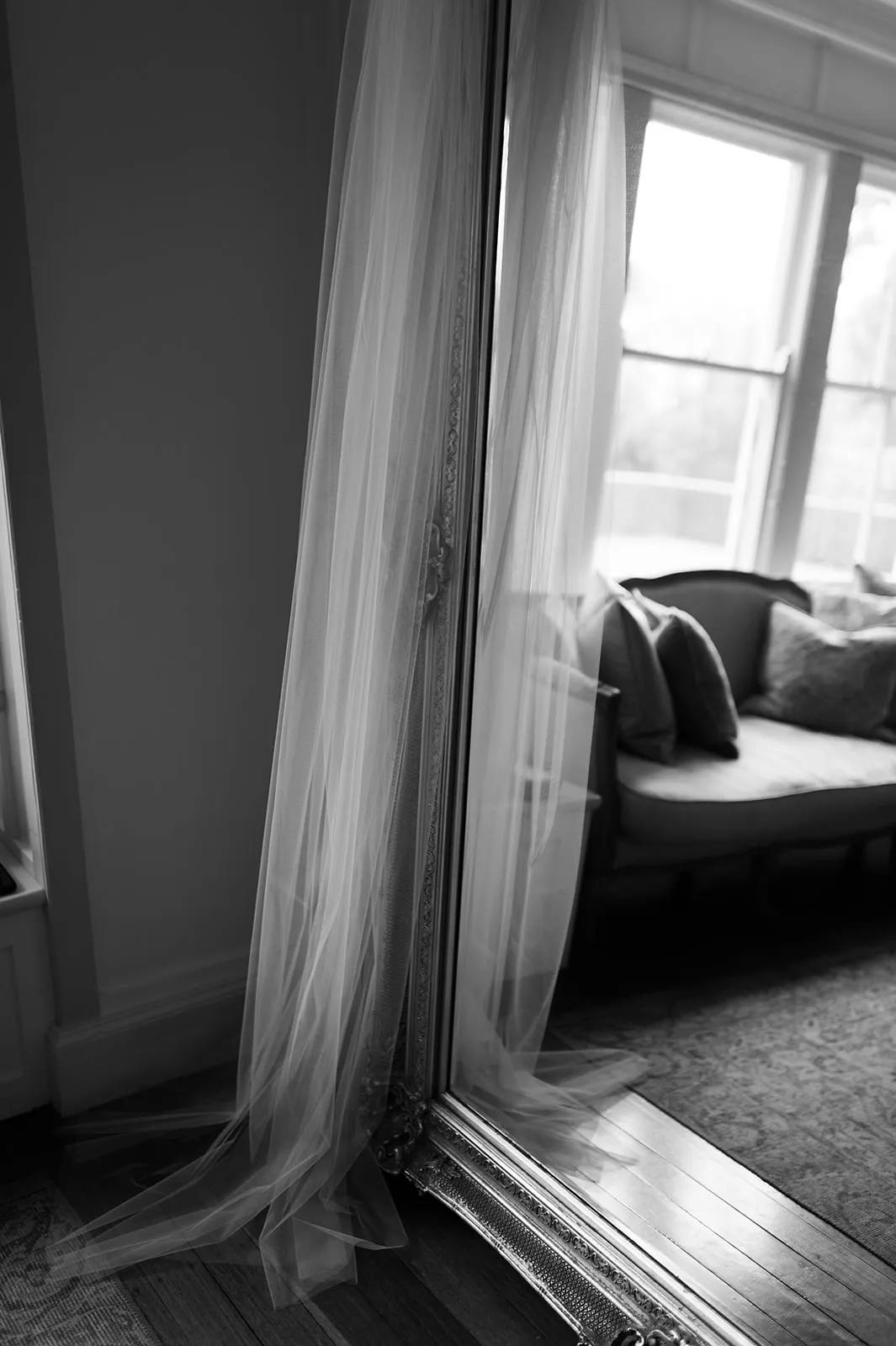 A long, sheer curtain lightly drapes over an ornate, full-length mirror leaning against the wall of a cozy room with a large window. In the background, a cushioned bench with throw pillows sits before the window, casting a soft, inviting ambiance.