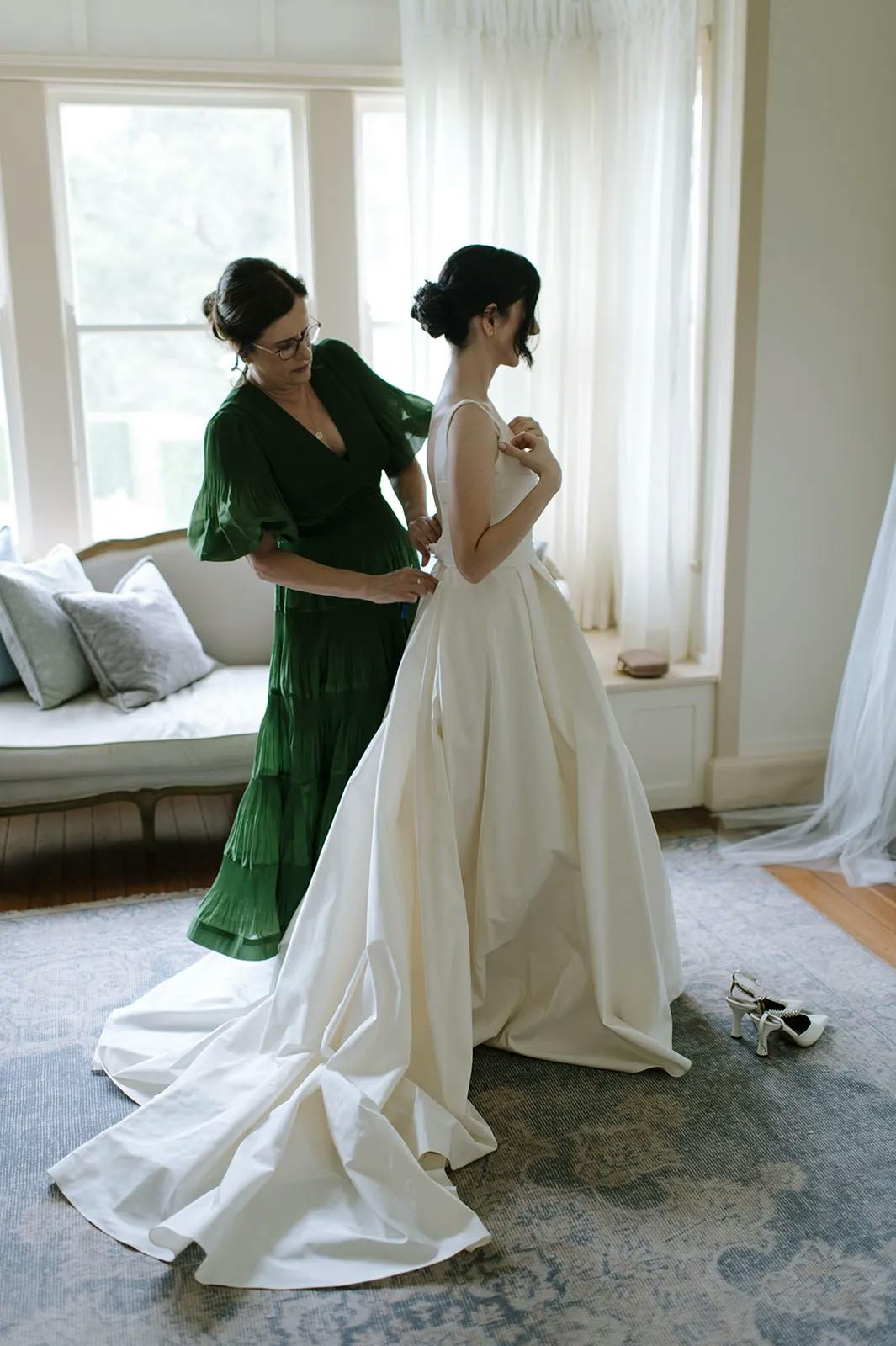 A woman in a white wedding dress stands while another woman in a green dress helps adjust the back of her dress. They are in a well-lit room with large windows, a couch with pillows, and a pair of high heels on the floor. The train of the wedding dress cascades behind.