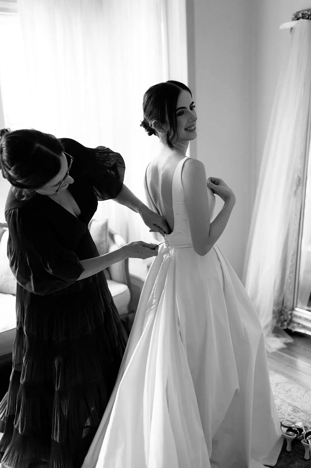 A bride, smiling and looking over her shoulder, stands in a room as another woman helps her with the back of her dress. The bride's gown is elegant with a flowing train. The scene is captured in black and white, adding a timeless quality to the moment.