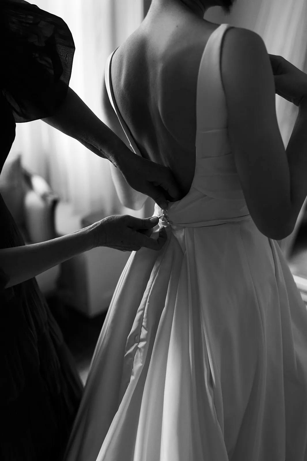 A black and white photo of a bride being helped by someone to fasten the back of her dress. The bride is wearing a sleeveless gown with an open back, and delicate hands are adjusting the fabric and buttons on her waist. The scene is set in a softly lit room.