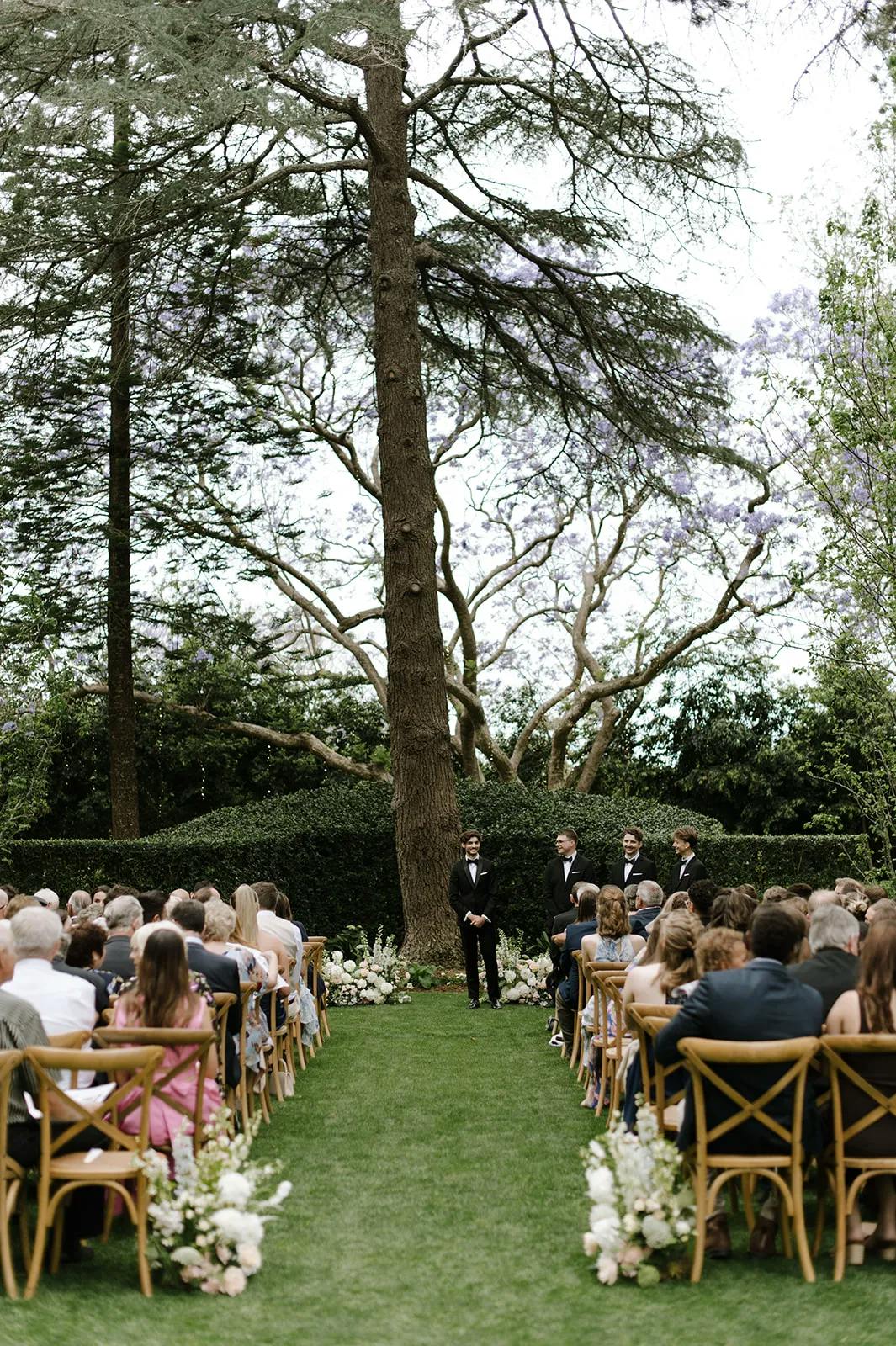 An outdoor wedding ceremony with guests seated on both sides of a grassy aisle, adorned with white flowers. The ceremony takes place under a large tree, where a groom and four groomsmen in suits stand at the end of the aisle, waiting.