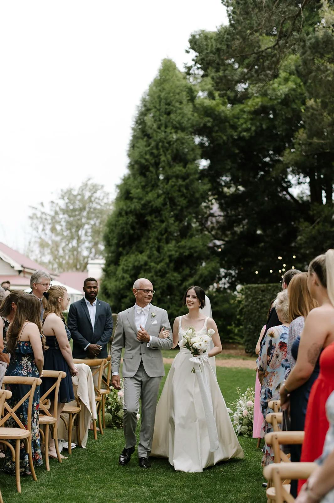 A bride, dressed in a white wedding gown and holding a bouquet, walks down an outdoor aisle with an older man in a light gray suit. Guests on either side of the aisle watch her, with greenery and trees in the background.