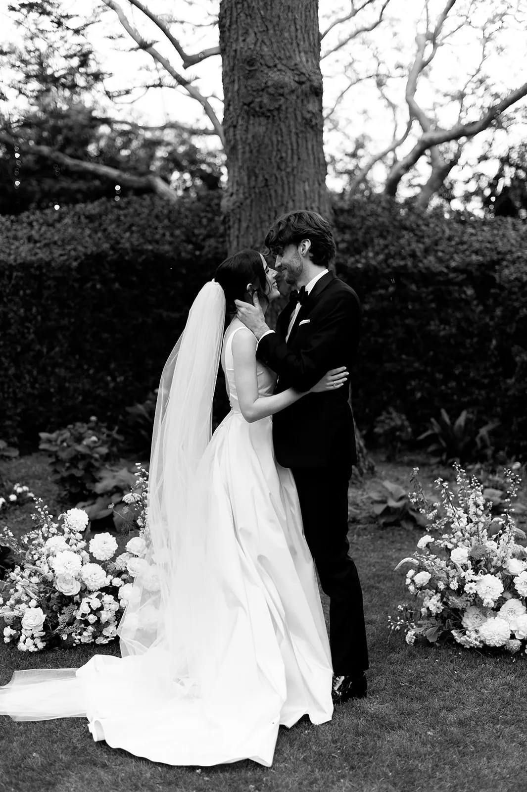 A bride and groom stand embracing in an outdoor setting. The bride wears a flowing white gown with a long veil, and the groom is dressed in a black tuxedo. They are surrounded by floral arrangements and greenery, with a large tree trunk in the background.