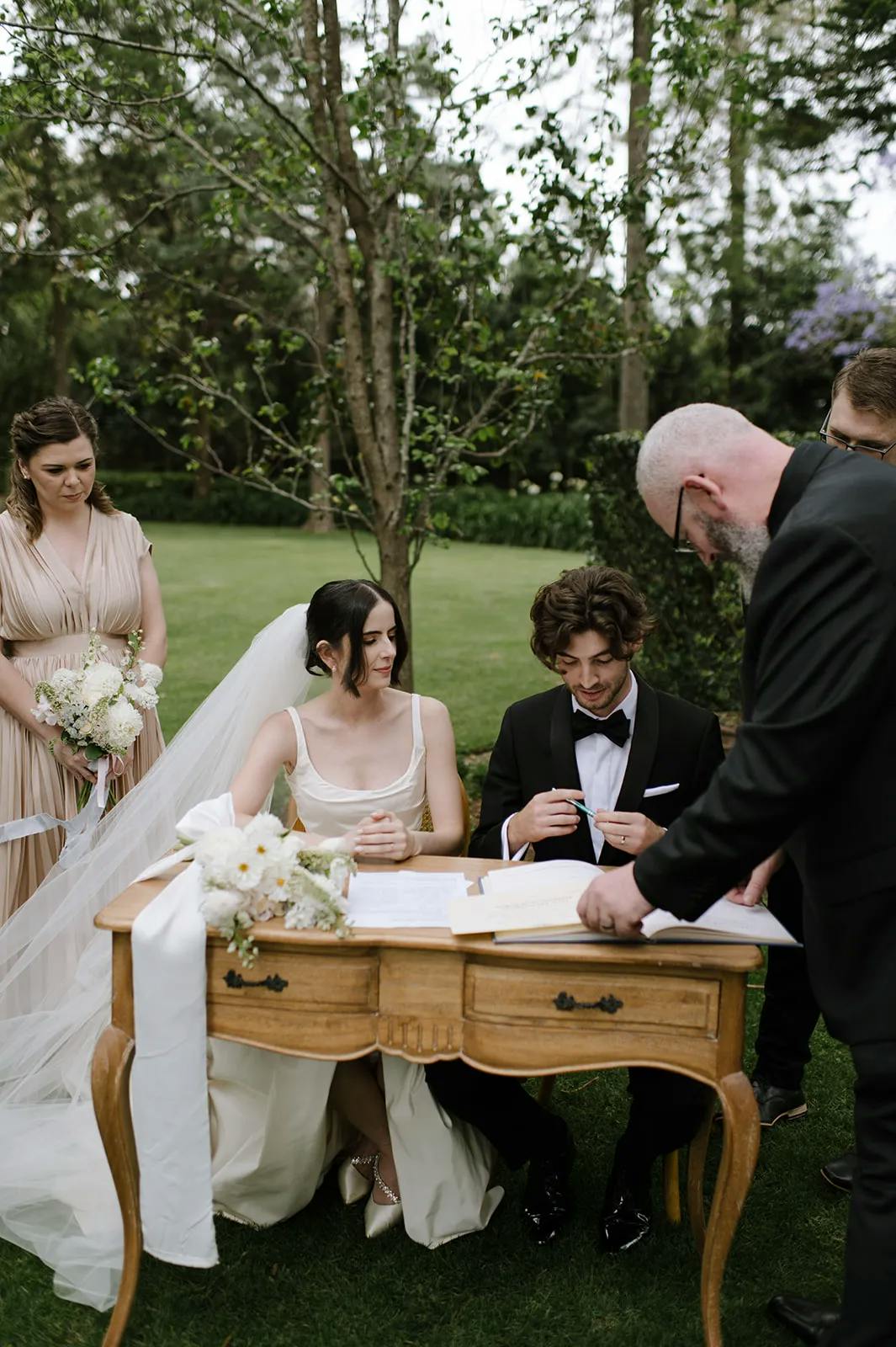 A wedding ceremony outdoors with a bride in a white dress and veil sitting at a wooden table, and a groom in a black suit sitting next to her. A bearded man is officiating. A bridesmaid in a beige dress stands holding flowers. Trees and greenery are in the background.