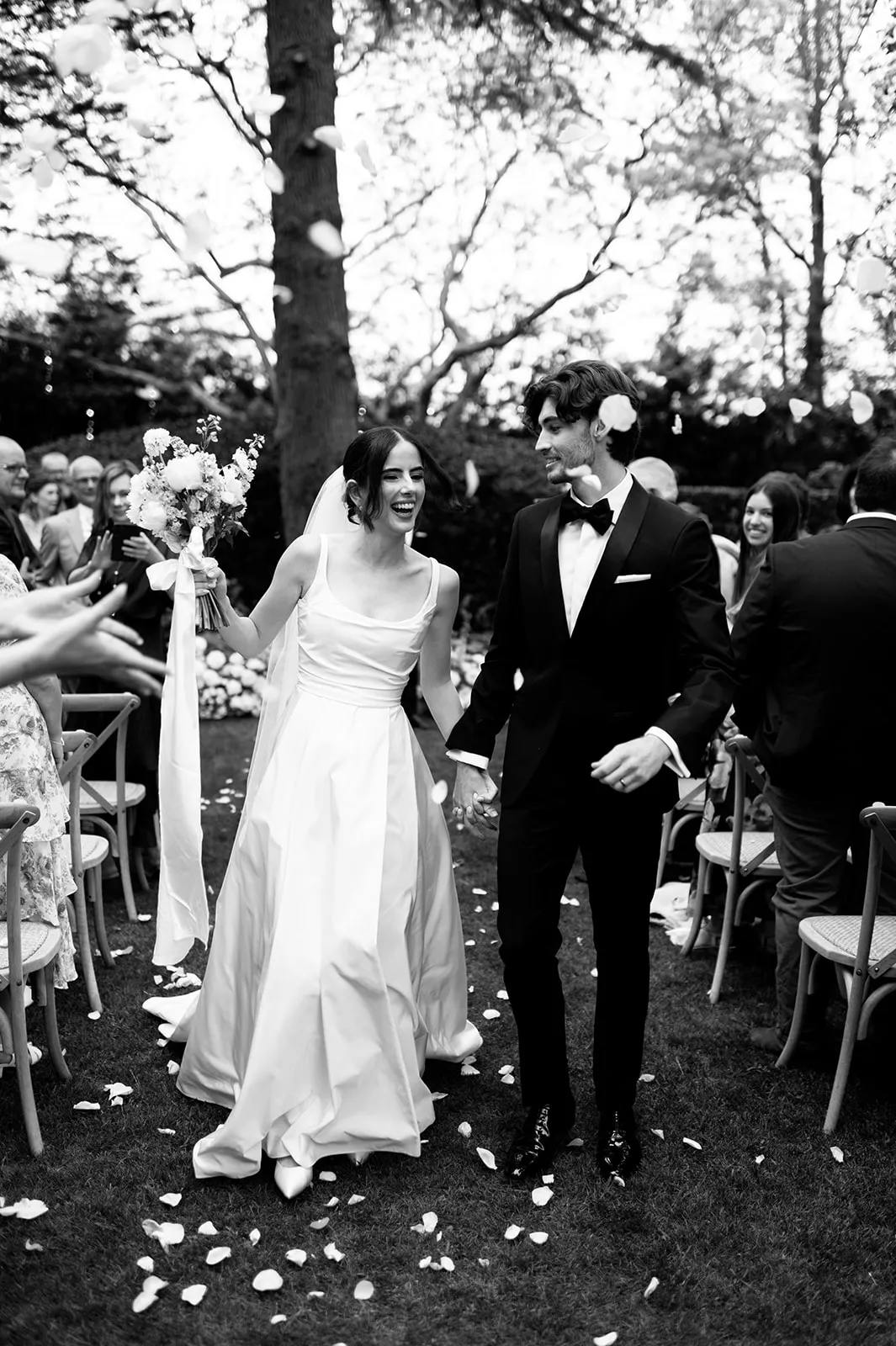 A bride and groom walk hand-in-hand down an outdoor aisle while guests cheer and throw flower petals. The bride is wearing a white wedding dress and holding a bouquet, and the groom is in a dark suit. The scene is set in a garden with trees in the background.