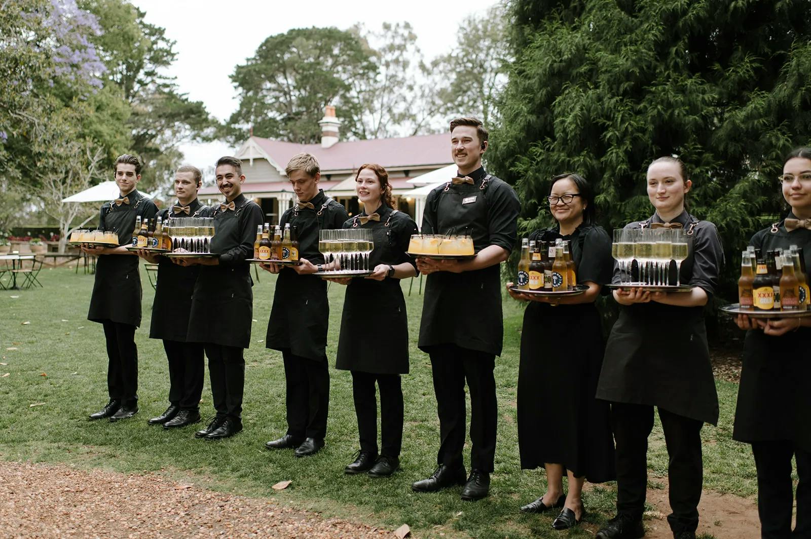 A group of nine servers, dressed in black uniforms, stand outdoors in a line holding trays with drinks and desserts. Behind them is a garden setting with trees, grass, and a building with a red roof. The atmosphere appears to be festive.