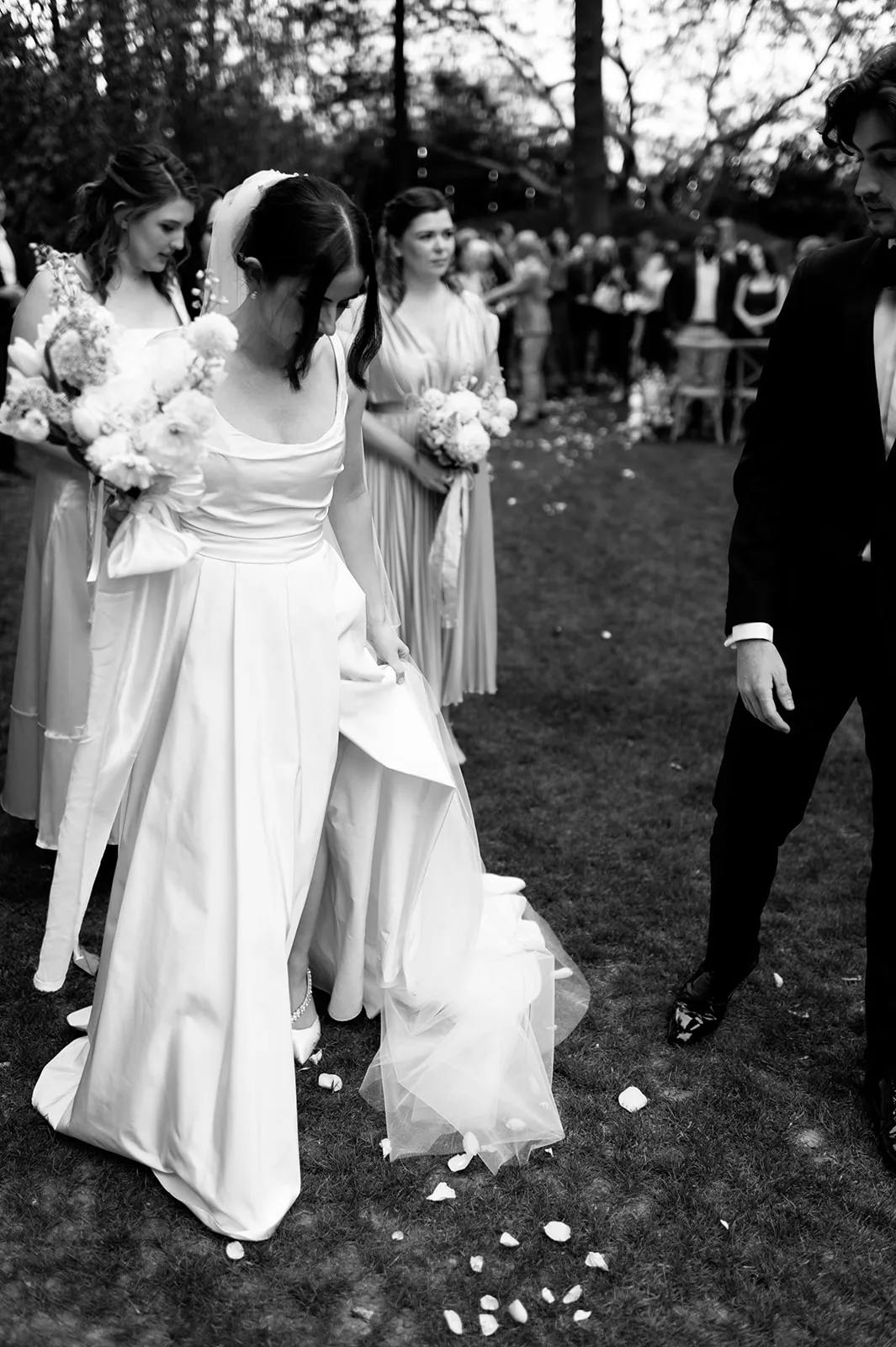 A black and white photo of a bride in a white gown holding her skirt, with bridesmaids behind her holding bouquets. A man in a tuxedo stands nearby. They are outdoors with blurred guests in the background. The ground is scattered with flower petals.