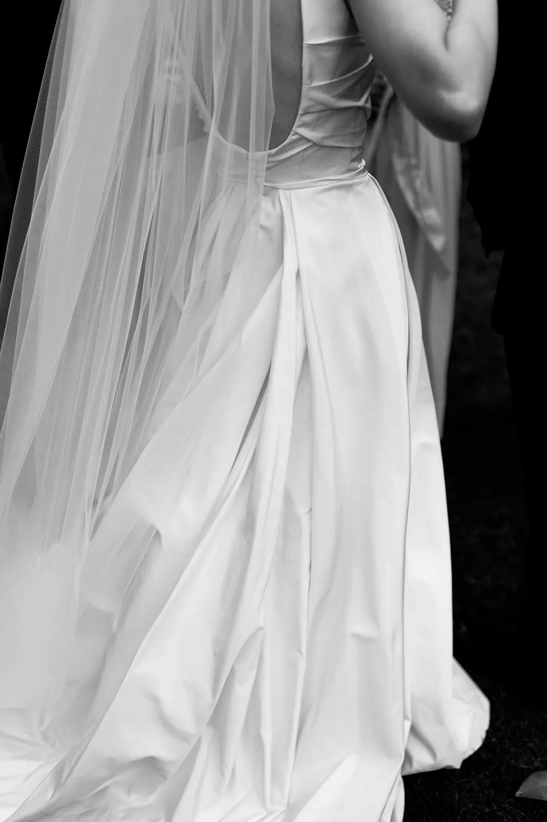 Black-and-white photo of a bride, focusing on the back of her wedding dress and veil. The dress features a low back and elegant folds, with the veil cascading down. Other people are slightly visible around her.